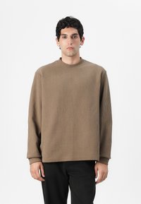 Top lungo a maniche lunghe di colore marrone con collo alto, vestibilità rilassata e spalle cadenti. La trama del tessuto sembra morbida e leggermente elasticizzata.