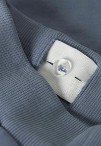 Una tela gris con textura acanalada presenta una etiqueta blanca con un botón transparente. El botón está ubicado en el centro, integrándose en el diseño.