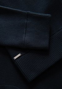 Dunkelblauer Strickpullover mit Rippbündchen und einem kleinen metallischen Etikett. Das Material ist weich und hat eine glatte Textur.