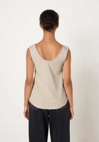 Tumejuustega naine, kes kannab beežikat varrukateta toppi ümarate seljaosaga ja musta laiade pükstega, seisab tagapool tavalise tausta ees.