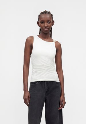 Femme aux cheveux tressés portant un haut blanc sans manches avec un encolure asymétrique et un pantalon noir taille haute sur un fond uni.