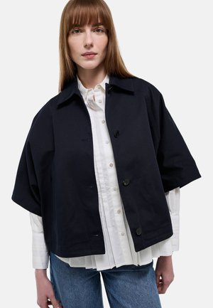 Jeune femme portant une veste noire ample boutonnée sur une chemise blanche à boutons et un jean bleu, debout devant un fond clair uni.