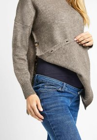 Pull en tricot marron clair avec un design large, présentant des boutons sur les côtés et des poignets côtelés, associé à un jean bleu foncé.