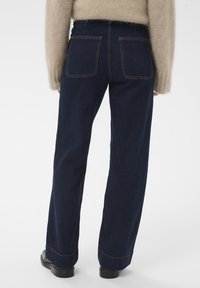 Mørkeblå denim bukser med bred ben, høj talje, to baglommer og gule syninger langs sømmene.