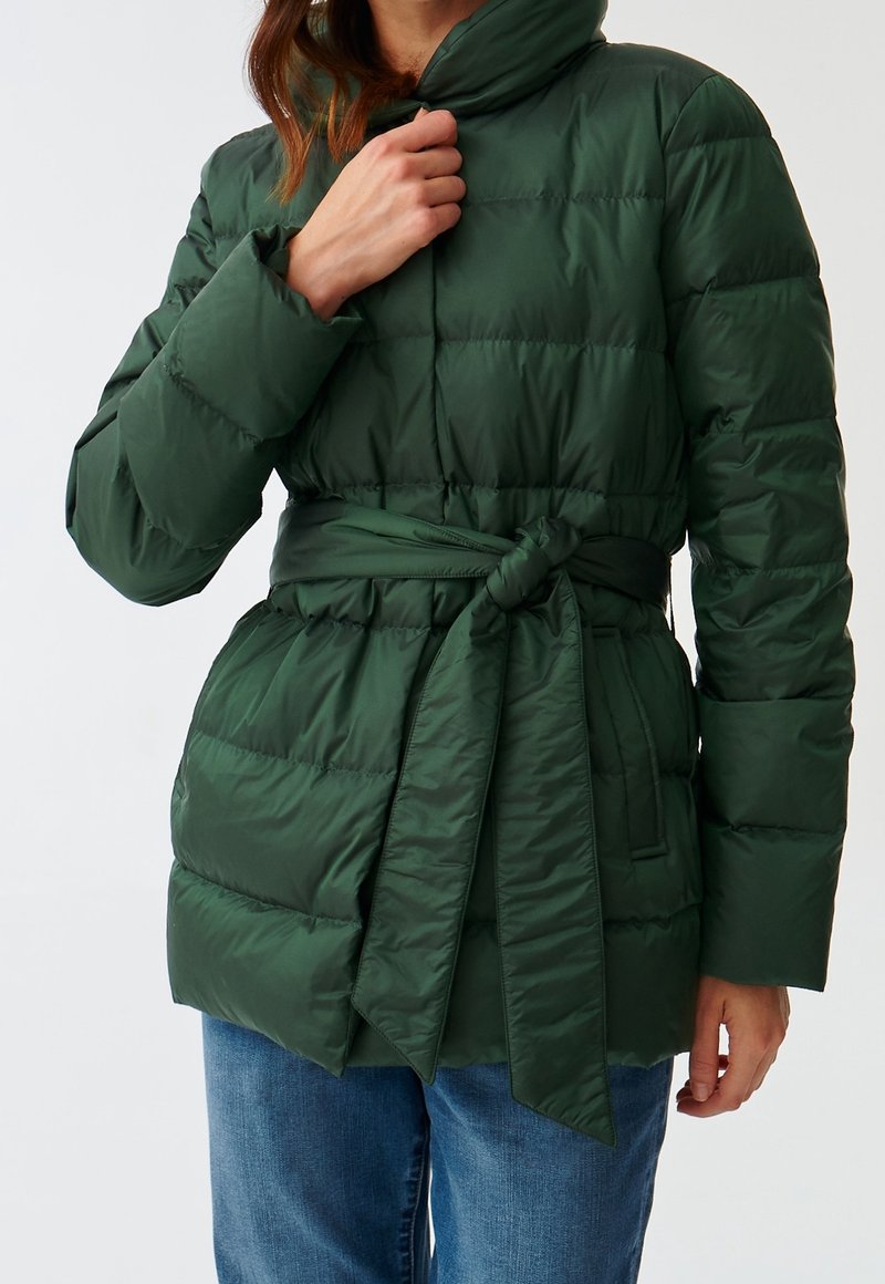Veste matelassée verte avec taille ceinturée, col montant et design matelassé. Caractérisée par un tissu lisse et une forme structurée, idéale pour le temps froid.