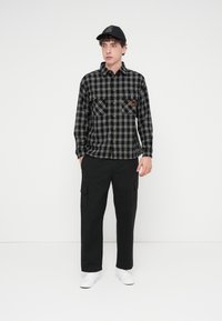 Καρό φανελένιο πουκάμισο σε μαύρο και άσπρο με δύο τσέπες στο στήθος, διακοσμητικά χρυσά κουμπιά, συνδυασμένο με μαύρο παντελόνι φορτών και λευκά αθλητικά παπούτσια.