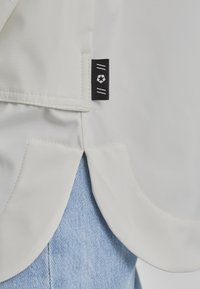 Helles beiges Gewebe mit glatter Textur, das eine Taschenapplikation und ein schwarzes Etikett mit weißem Sternembblem aufweist, über blauer Denim. 