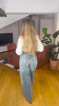 Svijetlosive traperice visokog struka s širokim nogavicama, s džepovima na leđima i izblijedjelom teksturom. Na sebi bijela majica dugih rukava.