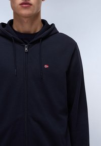 Felpa con zip di colore navy realizzata in tessuto morbido, con tasca frontale, cappuccio regolabile con lacci e un piccolo emblema con una bandiera sul petto.