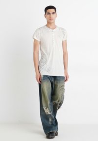 Vit kortärmad henley-tröja med knäppning fram, i kombination med storblå denimjeans med rivningar och en blekt grön nyans. Svarta stövlar.