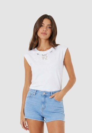 Donna con capelli castani lunghi indossa una canotta bianca senza maniche con decorazioni e pantaloncini di jeans azzurri, in piedi davanti a uno sfondo semplice.
