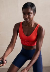 Red sports bra with cut-out detail and large "NIKE" logo on front, paired with fitted navy shorts, in a workout stance against a neutral background.