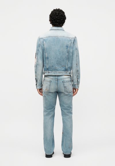 Άνδρας με σγουρά μαλλιά, που φοράει φθαρμένο ανοιχτό μπλε τζιν μπουφάν και αντίστοιχο τζιν παντελόνι, στέκεται με την πλάτη σε απλό φόντο.