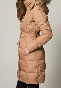 Abrigo de invierno acolchado beige con capucha, ribete de piel sintética, cintura ajustada y bolsillos laterales con cremallera; presenta un diseño acolchado y mangas largas.