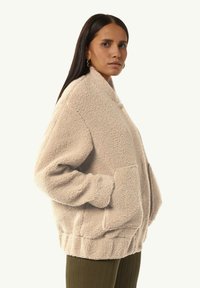 Beżowa, teksturowana kurtka polarowa z wysokim kołnierzem, bocznymi kieszeniami i ściągaczami, w połączeniu z oliwkowo-zielonymi spodniami z ribbanu.