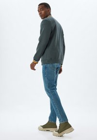 Suéter gris con puños a rayas amarillas y negras, combinado con jeans azules y botas verde oliva. El modelo está de pie mirando ligeramente hacia un lado.