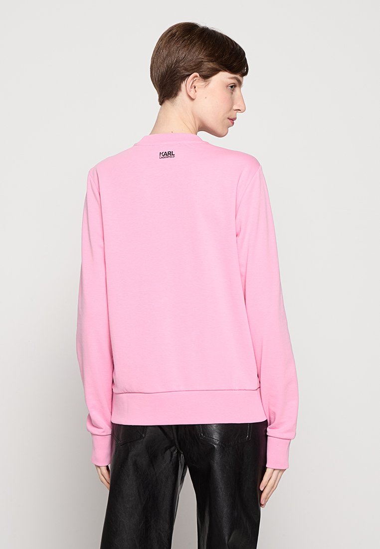 Rosa Sweatshirt mit langen Ärmeln, rundem Ausschnitt und gerippten Bündchen. Besitzt ein kleines schwarzes Logo auf der Rückseite. Getragen mit schwarzen Lederhosen.