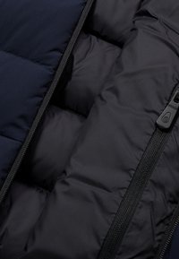 Mörkblå isolerad jackas interiör med svart foder, vaddering i kviltat mönster och en nylonblixtlås med detaljer i plast.