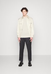 Camicia button-up beige chiaro sopra un maglione crema, abbinata a pantaloni scuri e scarpe nere. Design semplice con texture lisce.