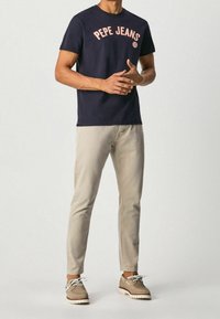 Marineblaues Baumwoll-T-Shirt mit „PEPE JEANS“-Logo in Zartrosa, kombiniert mit hellbeigen Hosen und hellbraunen Schuhen, die einen lässigen Look präsentieren.