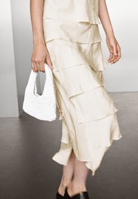 Beige gestuftes Kleid aus strukturiertem Stoff, begleitet von einer weißen geflochtenen Handtasche. Das Kleid hat geschichtete Rüschen und einen schrägen Saum.