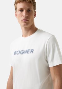 Jonge man met krullend blond haar, gekleed in een wit T-shirt met vervaagde 'BOGNER'-tekst aan de voorkant, staat rechtop tegen een effen achtergrond.