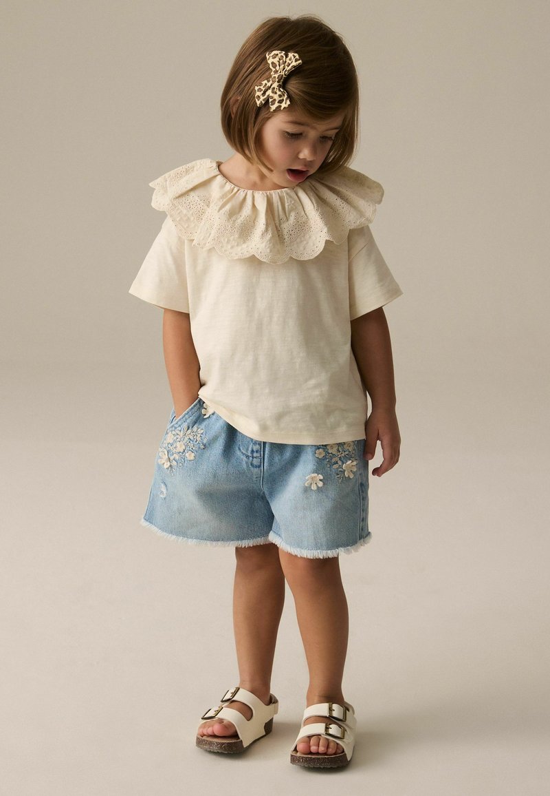 Jeune fille portant un haut crème à col à volants, un short en jean avec broderies florales, des sandales blanches et un nœud papillon pour cheveux imprimé léopard, regardant vers le bas.