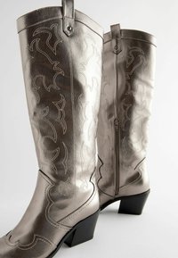 Metalliset hopeiset cowboy-saappaat koristeellisilla ompeleilla, polven keskikorkeus, vetoketjukiinnitys ja musta kerroksellinen korko. Sujuva tekstuuripinta.