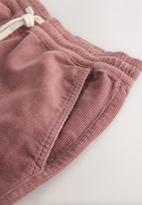 Gros plan d’un pantalon en velours côtelé rose poussiéreux avec taille élastique, poche latérale et cordon blanc sur fond clair.