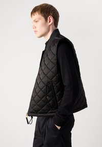 Black quilted vest with a high collar, zip closure, and side pockets. Features a smooth texture and contrasting dark sweater underneath.