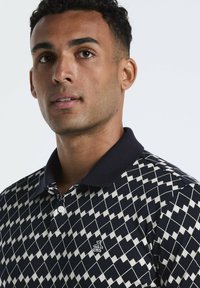 Polo shirt à motifs marine et crème avec des formes de losanges, manches courtes et col classique. Présente un accent de logo sur la poitrine.