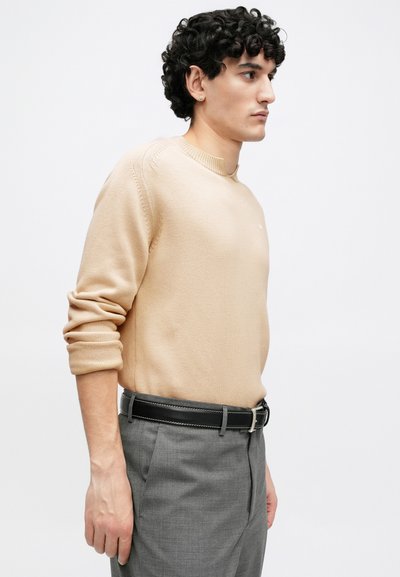 Homme aux cheveux bouclés portant un pull beige aux manches retroussées, un pantalon gris et une ceinture noire, debout de profil sur un fond blanc.