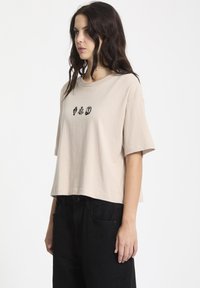 Beige lyhyt t-paita pehmeästä puuvillasta, jossa on pyöreä pääntie ja lyhyet hihat. Edessä musta graafinen painatus, jossa on erilaisia motivoita.