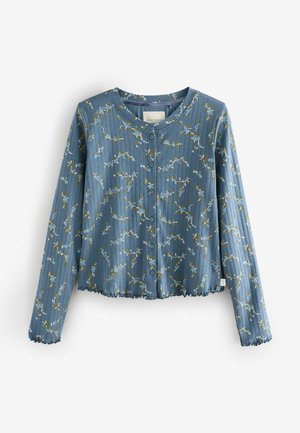 Mėlynas šonkačiuotas megztinis su gėlių raštu, apvaliu kaklu, mygtukais priekyje ir volanėlėmis ant hem ir rankovių. Pagamintas iš minkštos, lengvos medžiagos.