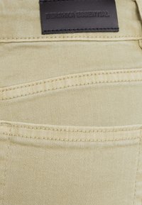 Tejido de mezclilla beige con una textura sutil. Cuenta con bolsillos y una etiqueta de cuero negro con "BERSHKA ESSENTIAL" grabada.