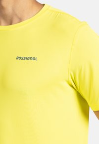 Klart gul kortärmad träningsskjorta gjord av ett mjukt, lätt material med rund halsringning och en kontrasterande grå logotyp på bröstet.