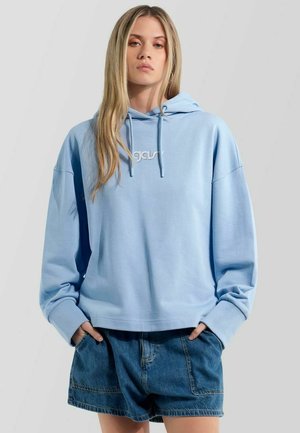 Giovane donna con felpa con cappuccio azzurra chiara e shorts di denim larghi con le mani nelle tasche, su uno sfondo chiaro e semplice.