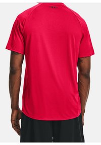 Muž v jednobarevném červeném sportovním tričku s krátkým rukávem a malým černým logem Under Armour na horní části zad, doplněný o černé šortky.