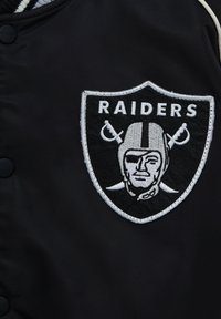 Zwarte satijnen jas met een witte geborduurde Raiders-logo. Het logo bevat een met helm geklede figuur geflankeerd door gekruiste zwaarden.