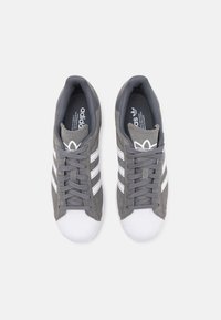 Šedé semišové sportovní boty s bílou gumovou špičkou a třemi bílými pruhy po stranách. Mají polstrovaný šedý interiér a tkaničky.