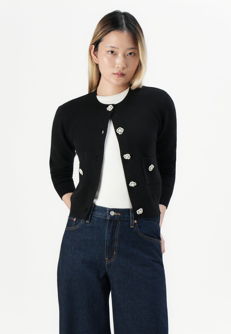 Jeune femme aux cheveux raides portant un cardigan noir avec des boutons décoratifs sur un haut blanc et un jean taille haute bleu foncé, les mains dans les poches.