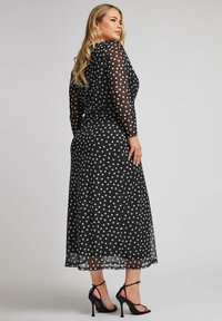 Mustaläiskukka-mekko, jossa on pitkät läpinäkyvät hihansuut, midi-pituus, joustava vartaloosa ja laskostettu hame. Pukeutuneena mustiin nauhakenkiin.