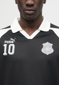 Moški, ki nosi črno športno majico z belim ovratnikom, logotipom Puma, številko 10 na levi strani prsnega koša in zapletenim dizajnom Puma ščita na desni strani prsnega koša.