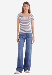 Gestreept T-shirt met korte mouwen, wit met marineblauwe strepen, gecombineerd met wijde blauwe jeans. Model draagt lichtgekleurde sneakers en staat rechtop.