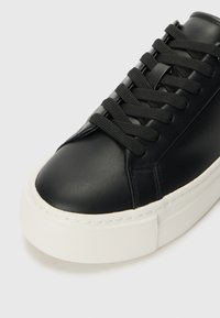Basket en cuir noir avec un bout rond, surface texturée et semelle en caoutchouc blanche ; elle présente des lacets plats noirs et des détails de couture minimalistes.