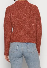 Rustfarbener, gestrickter Pullover mit strukturiertem Finish, Rundhalsausschnitt und leicht verkürztem Design. Kombiniert mit hellblauen Jeans.