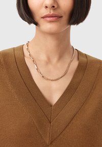 Maglione marrone a maglia con scollo a V e dettagli testurizzati, abbinato a una collana con catena in oro che presenta anelli lisci e testurizzati.