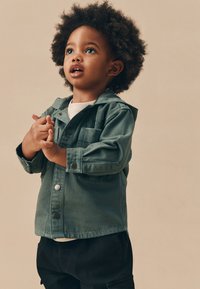 Jachetă verde din denim cu glugă, buzunar la piept și nasturi tip capse. Purta peste o cămașă albă, asortată cu pantaloni negri. Păr creț.