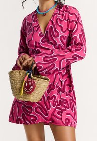 Femme en robe portefeuille rose à motifs tenant un sac tissé orné d'un porte-clés visage souriant, portant des colliers de perles bleues et roses superposés.