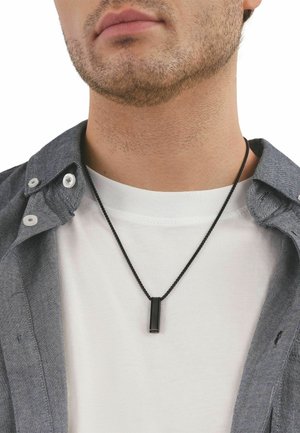 Joven con barba corta que lleva una camiseta blanca, una camisa azul abierta y un colgante rectangular negro en un collar de cordón negro.